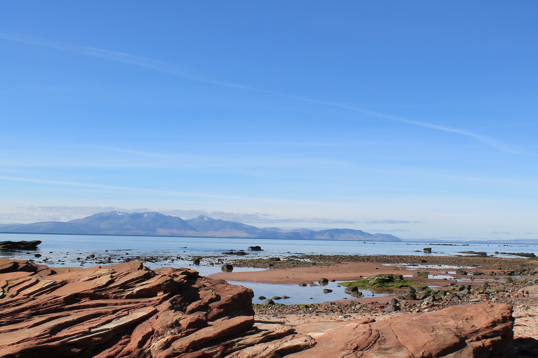 In the foreground is a redish brown rocky outcrop.  Beyond is a stony and sandy beach. Across the water, land with large hills can be seen in the distance.