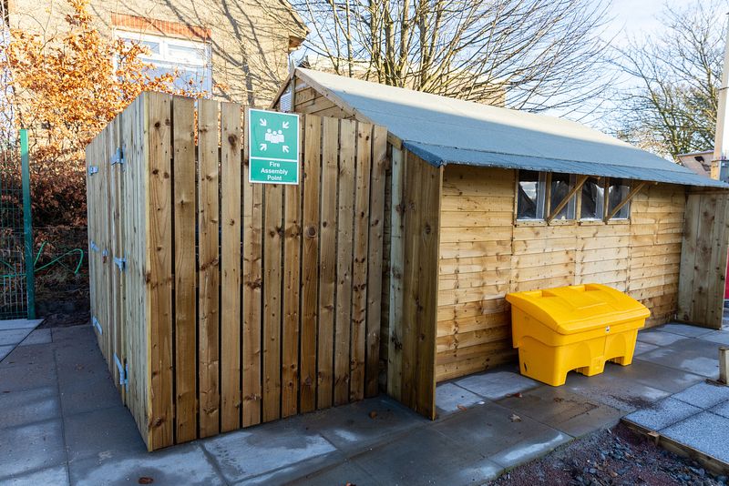 A wooden building with a gently sloped roof. To the left is a fenced off area with a green sign saying 'fire assembly point' attached to the tall wooden fencing.