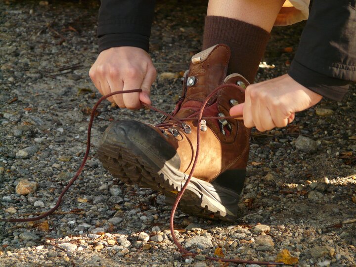 We see someone wearing sturdy brown walking shoes on a gravel path in the sunlight. They are grasping the laces and tying them.