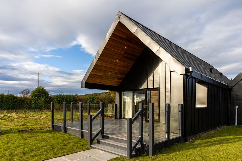 Todhill chalets in the sunshine. They have a black exterior with wide decking and french doors.