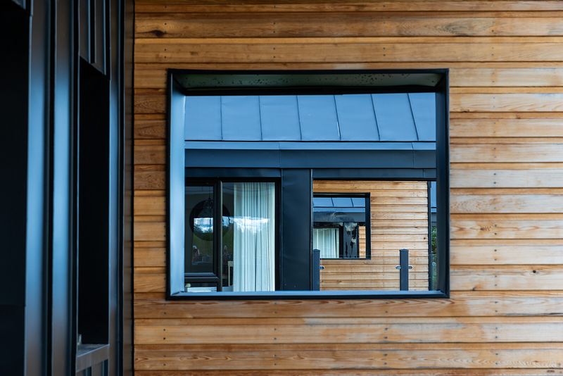 A window is in the wooden panelled exterior of the chalet, in the reflection we can see other chalets behind.