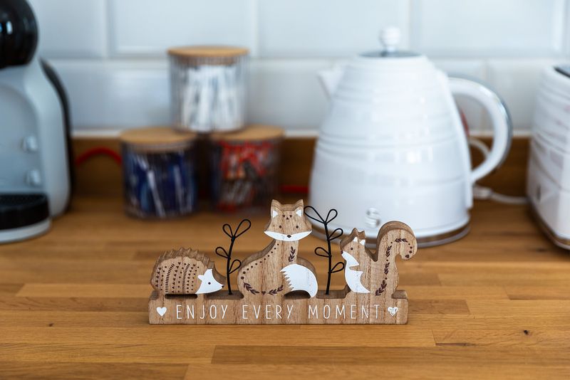 A charming wooden ornament stands on the surface in front of a kettle and other kitchen items. It says 'enjoy every moment' and features little woodland animals.