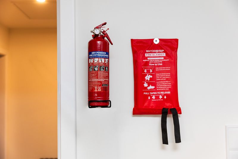 A red fire extinguisher and red fire blanket are attached side by side to a white wall. The extinguisher says 'dry powder fire extinguisher' at the top against a blue background. The blanket has 'how to use' text.