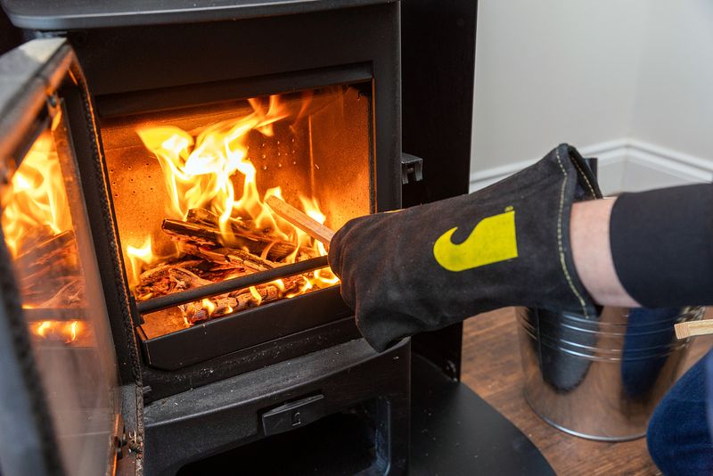 The furnace is lit and open with small pieces of wood burning fully. A gloved hand is handling a piece of wood near the flame.