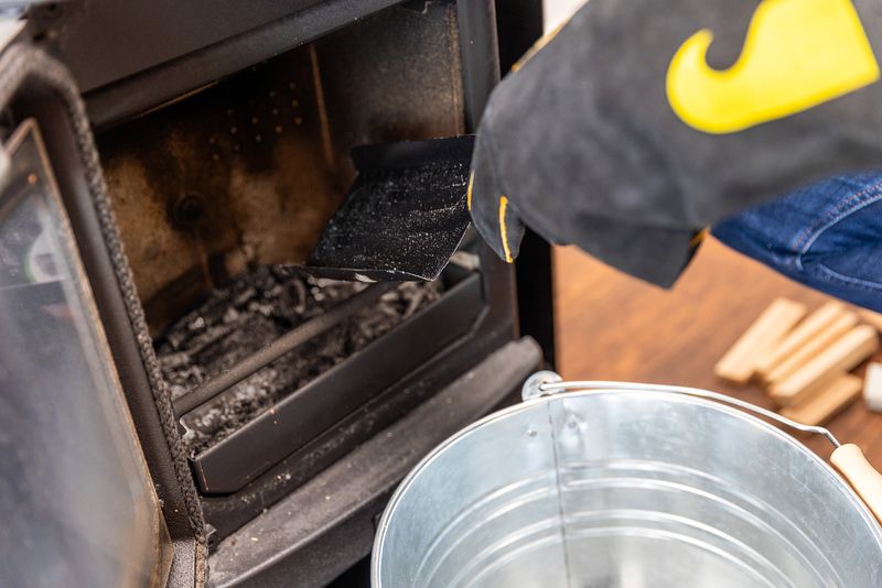 The stove is unlit and open. It is filled with remnants from the fire. A gloved hand is holding a small shovel like tool.