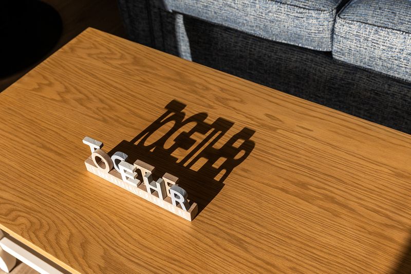 A small wooden sign with the word 'together' sits on a table in the daylight. It is casting a soft shadow which also says 'together'.