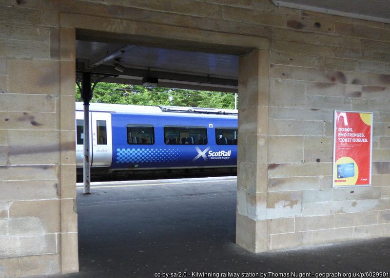 A blue train can be seen through an archway in a stone wall at the train station.